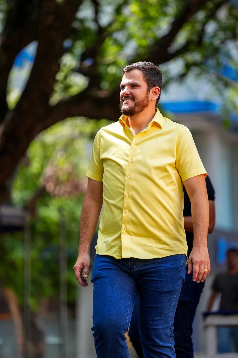 Renato Junior, de camisa amarela, caminha sorrindo sob uma árvore